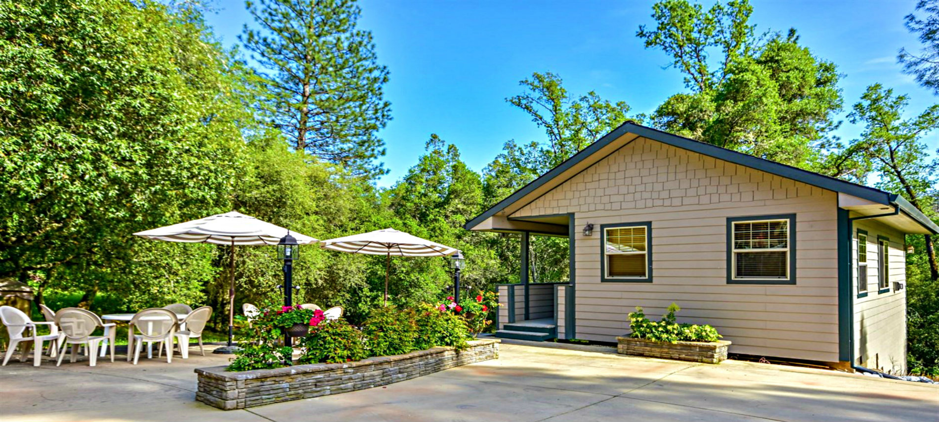 A yellow cottage with blue trim next to a patio with a stand of green trees behind it.