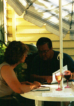 A man and woman at a patio table with umbrella and working material on the table with wine glasses.