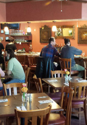 The inside of a restaurant with two men at a wine bar and people sitting at dining tables